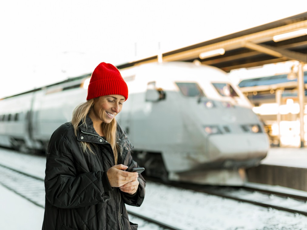 En kvinna med röd mössa står vid en tågperrong framför ett tåg medan hon tittar på sin telefon.